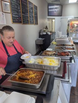 an older woman with greying dark hair pulled back into a bun serves meat and veggies in thick colorful sauces from big roasting pans