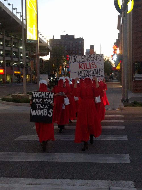 a group of people in red Handmaid's Tale costumes cross a street holding songs such as 'Fortenberry Kills Nebraskans'