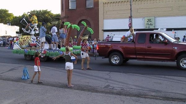a red truck pulls a float labeled C&C Beach Party. There are lots of people on or around it, and a guy in a Hawaiian shirt about to launch a hot dog from the slingshot seat