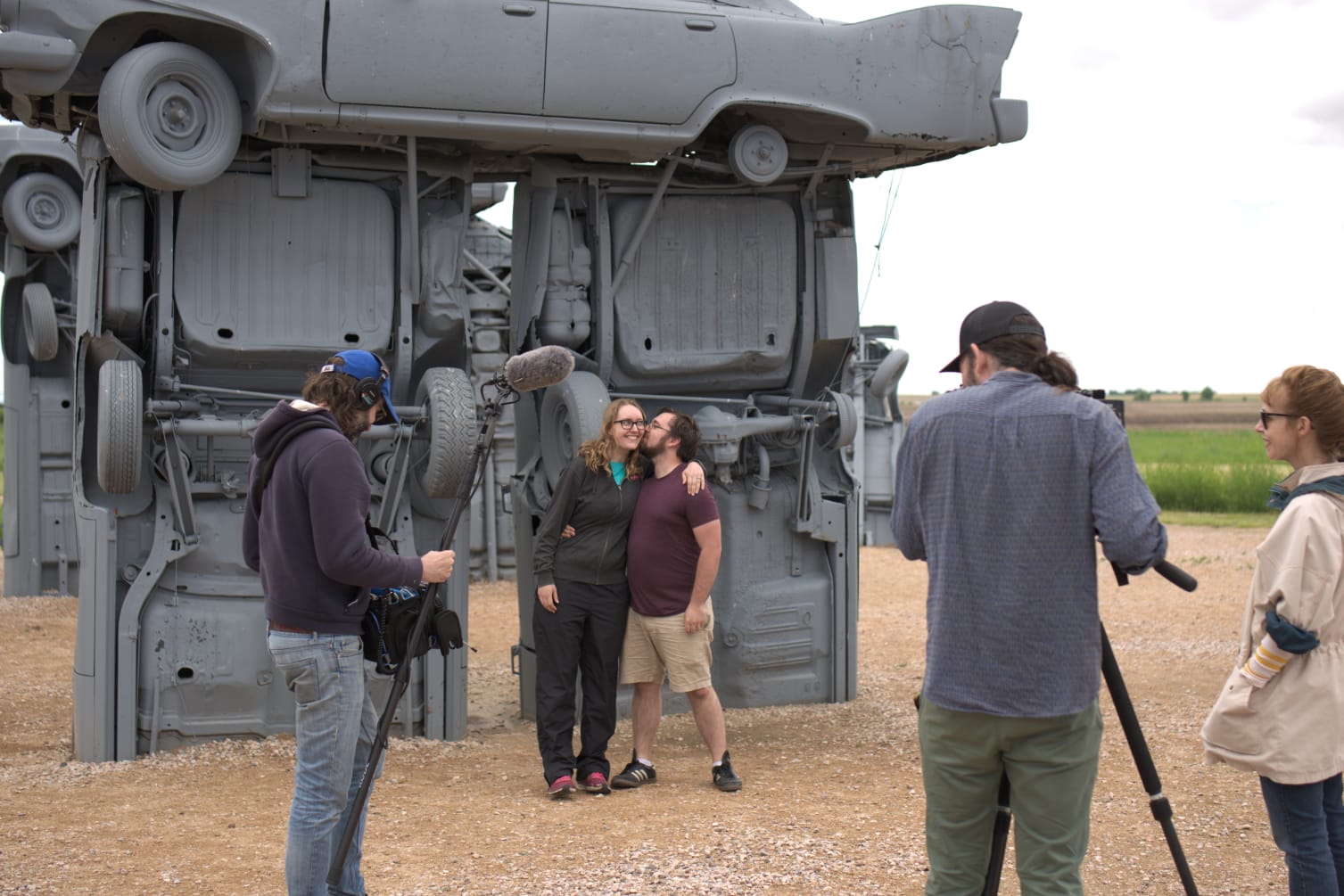 Wren and Cassey in front of some grey-painted cars stood up on their bumpers, surrounded by a camera crew