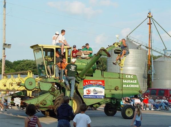 a green combine has cut-out pig shapes along the rotating head in front, and the hot dog slingshot thing mounted on back. lots of people are standing in other places on the combine